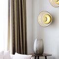 Round glass wall light lit on a pale corner wall, beside a linen curtain and a ribbed side table.