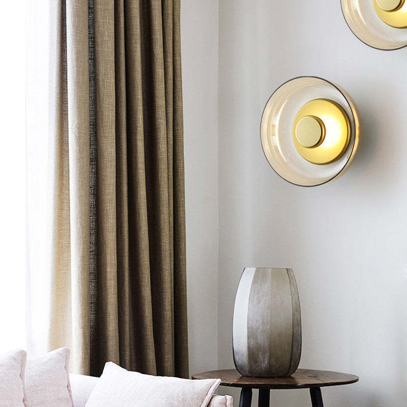 Round glass wall light lit on a pale corner wall, beside a linen curtain and a ribbed side table.