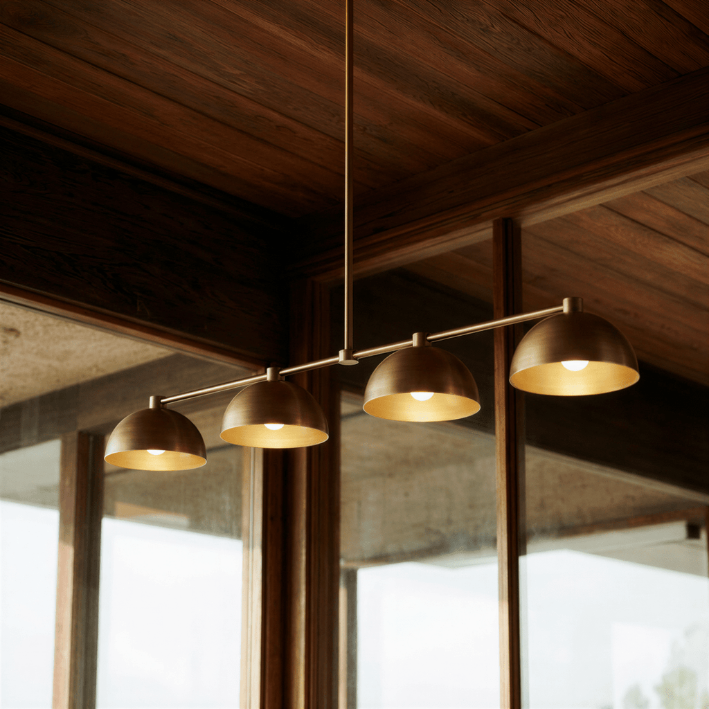 Clérachic four-dome bar pendant light in aged brass, seen close from below with a lamp showing inside each open dome, under a dark timber ceiling.