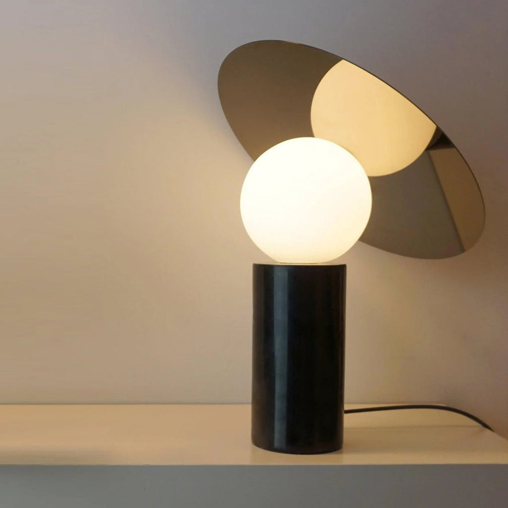 Against a taupe wall, the Clérachic disc lamp glows on a bedside table, its black marble base standing on a dark wood cabinet.