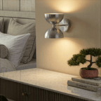 Clérachic brushed chrome double-dome wall sconce lit above a credenza with a bonsai tree and stacked books.