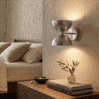 Clérachic brushed chrome double-dome wall sconce lit above a bedside table with a vase and stacked books.