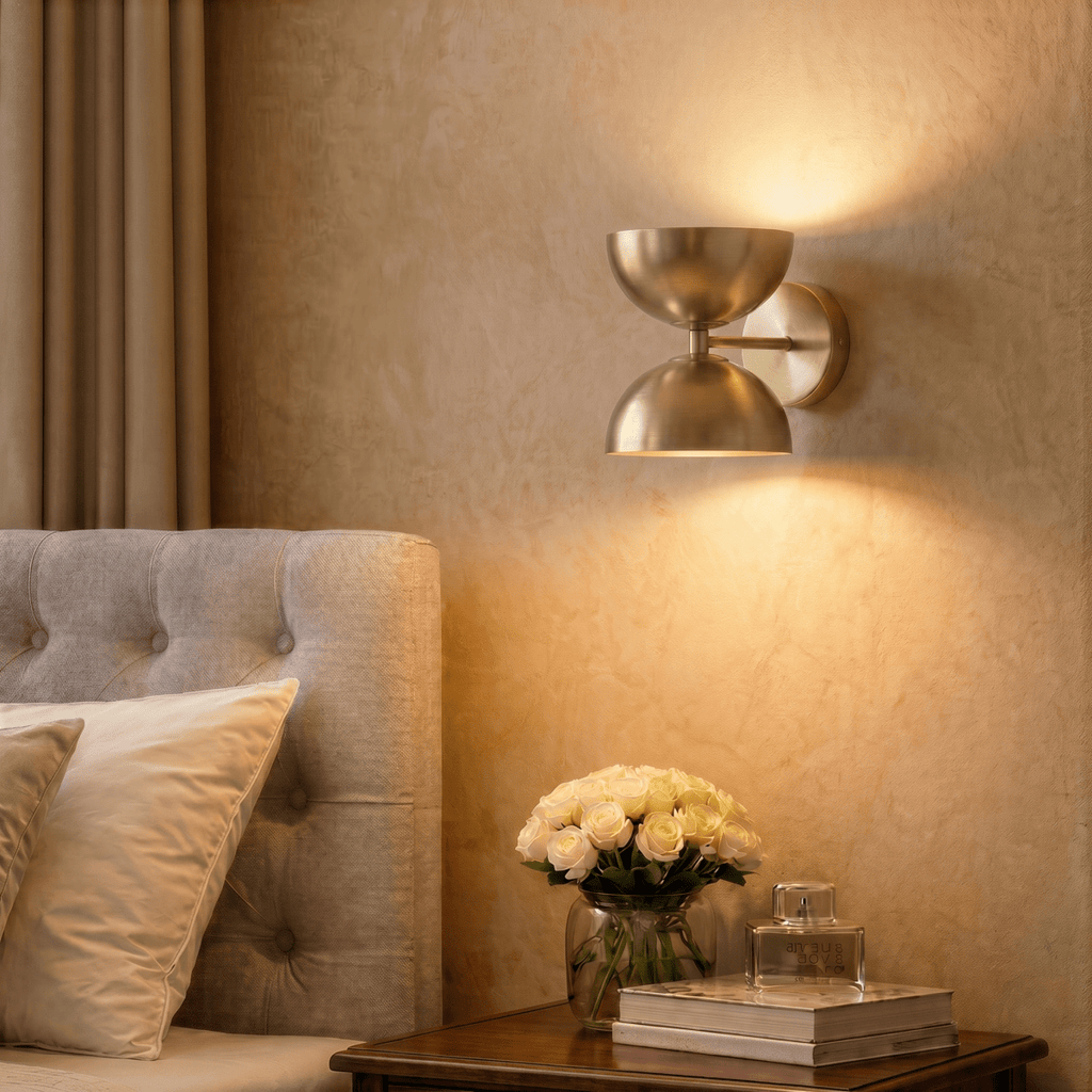 Clérachic polished brass double-dome wall sconce lit above a bedside table with white roses and a perfume bottle.