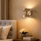 Clérachic polished brass double-dome wall sconce lit above a bedside table with white roses and a perfume bottle.