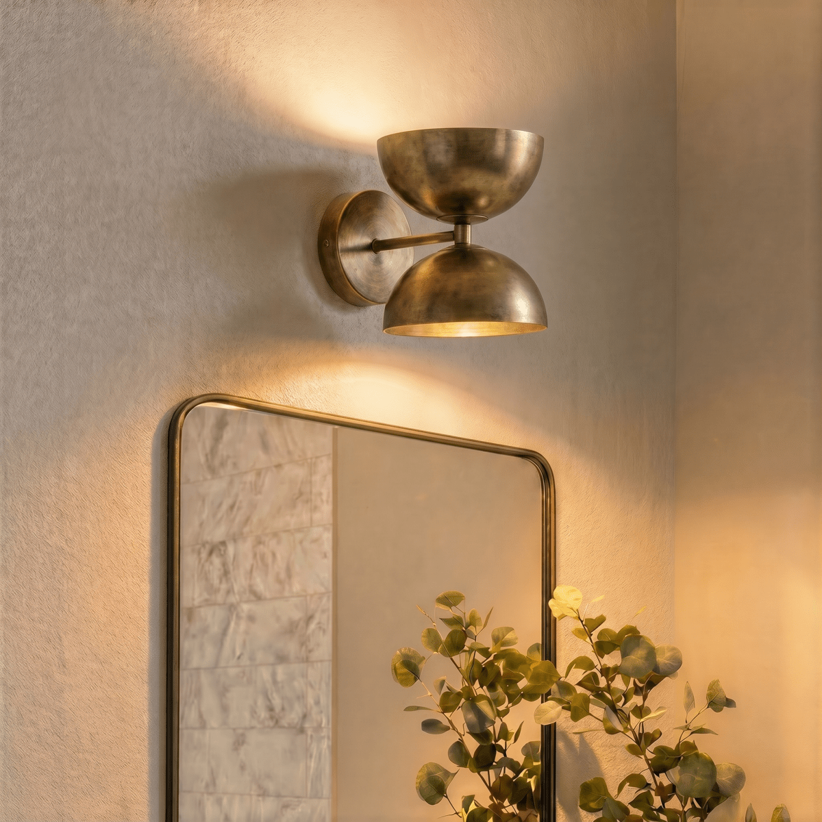 Clérachic vintage brass double-dome wall sconce lit above a framed mirror beside a leafy eucalyptus branch.