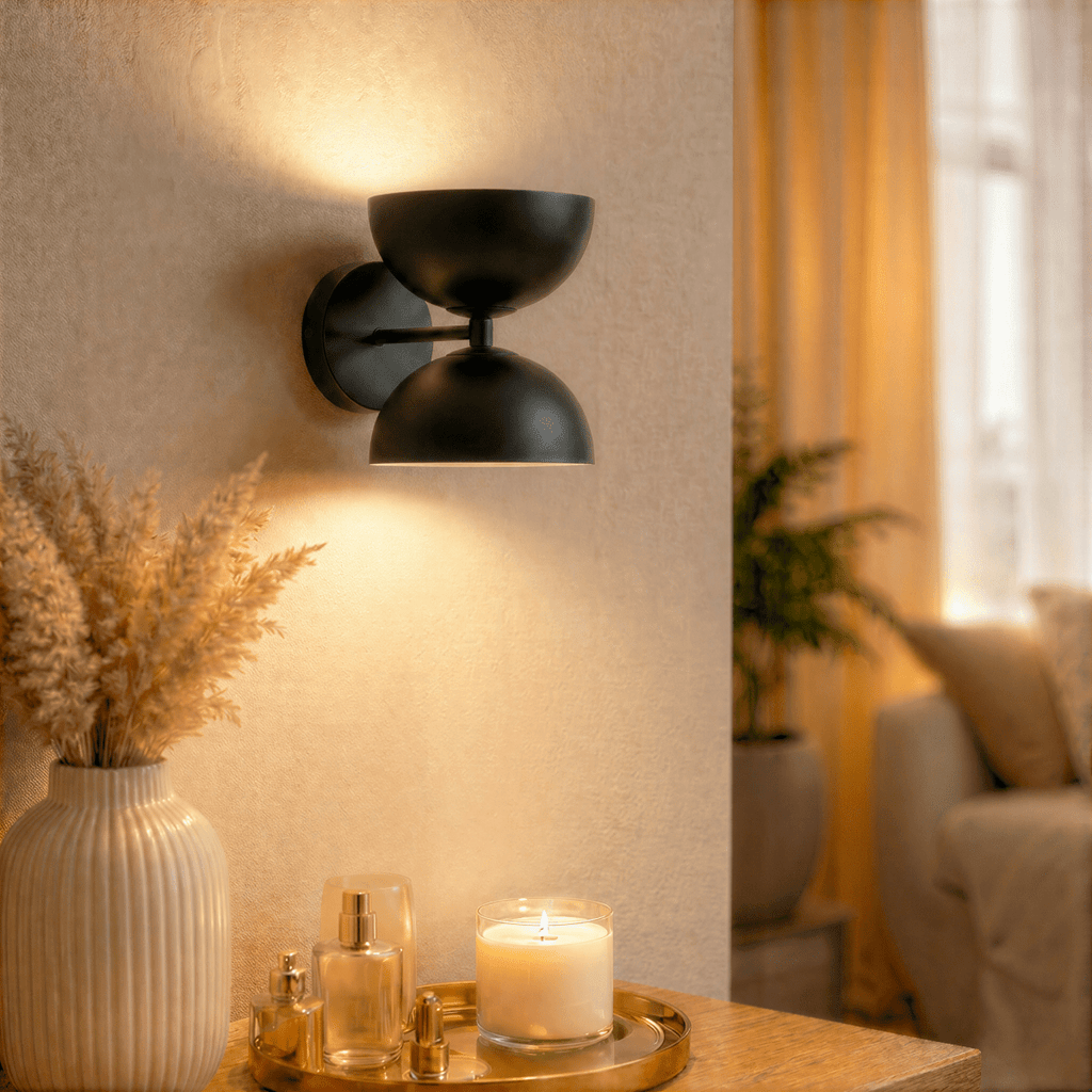 Clérachic matte black double-dome wall sconce lit above a console table with a vase, candle and perfume bottles.