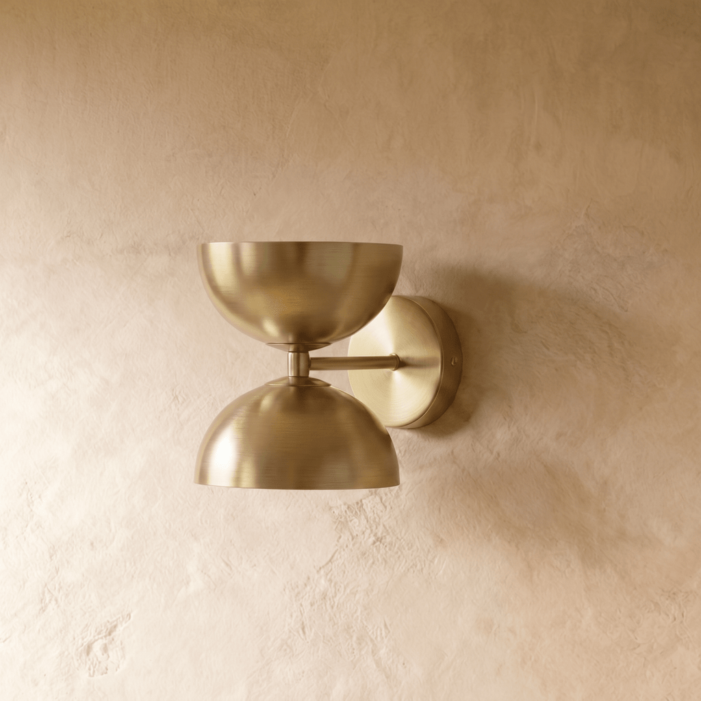 Clérachic double-dome wall sconce in polished brass, shown closer-cropped against a plain plaster studio wall.