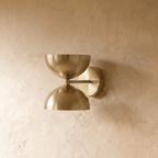 Clérachic double-dome wall sconce in polished brass, shown closer-cropped against a plain plaster studio wall.