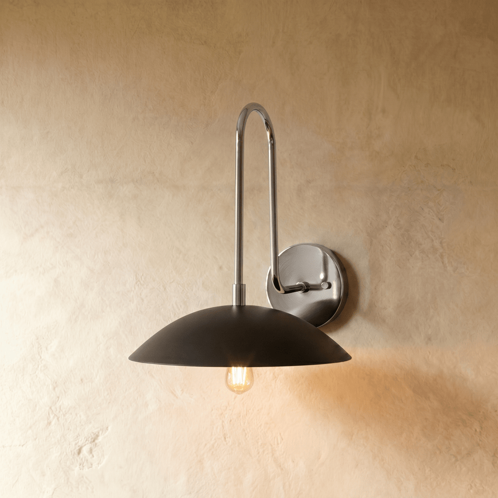 Clérachic wall lamp in chrome with a black dome shade, the gooseneck arm catching light against a plaster wall.