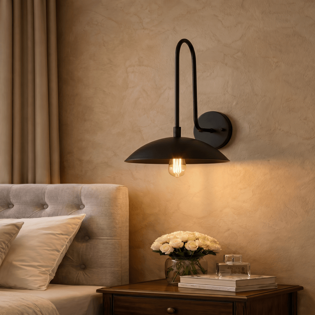 A black Clérachic wall lamp lights a bedroom nightstand holding white roses and a small stack of books.