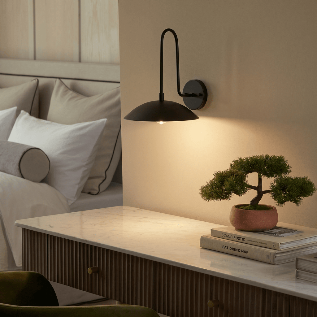 A black Clérachic wall lamp lights a marble-top dresser holding a small potted bonsai and stacked books.