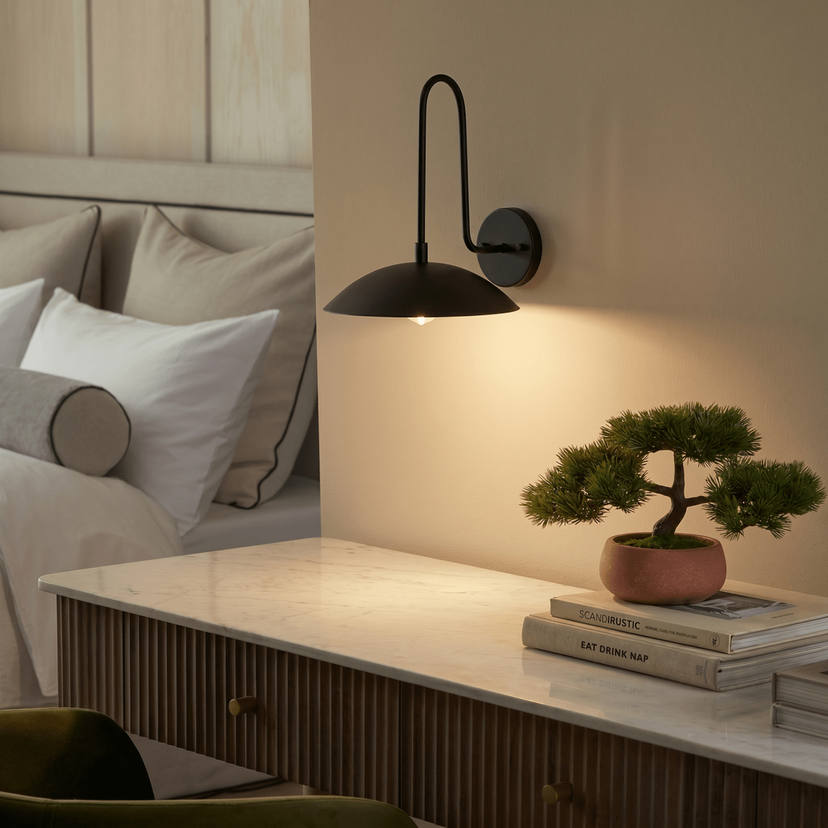 A black Clérachic wall lamp lights a marble-top dresser holding a small potted bonsai and stacked books.