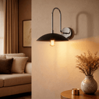 A chrome Clérachic wall lamp with a black dome shade lights a living room corner near a patterned vase.