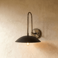Clérachic wall lamp in vintage brass with a matte black dome shade, its bulb glowing softly against a plaster wall.