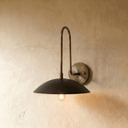 Clérachic wall lamp in vintage brass with a matte black dome shade, its bulb glowing softly against a plaster wall.