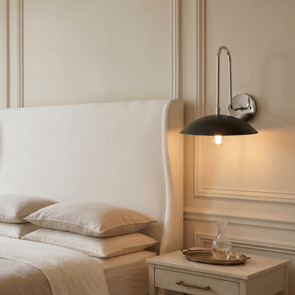 A chrome Clérachic wall lamp lights a bedroom nightstand holding a glass carafe on a tray, beside an upholstered headboard.