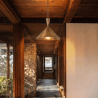 Clérachic cone-shade pendant light hanging from a timber ceiling at the start of a stone-and-timber hallway, lit, with a sunlit doorway at the far end.