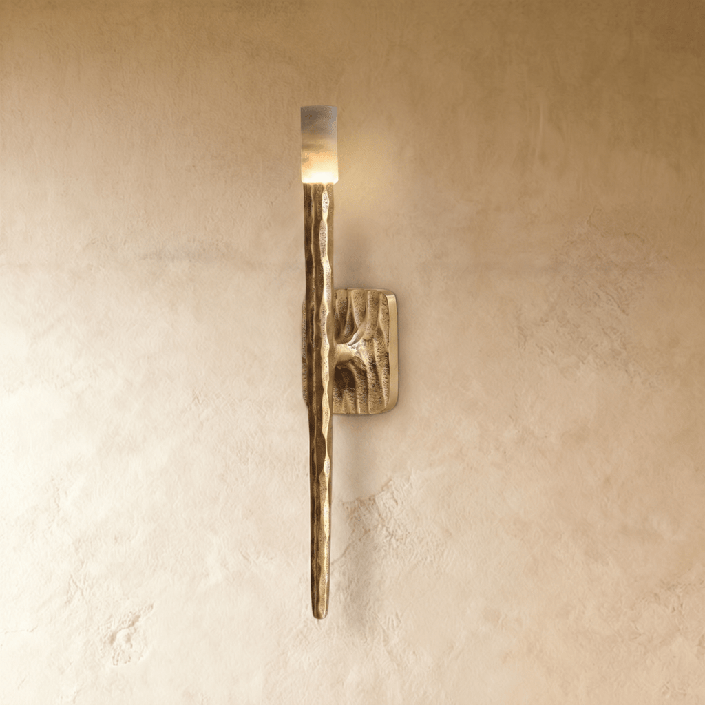 Clérachic wall lamp lit on a plaster wall, its pale stone cap glowing above a ridged copper-toned blade that narrows to a fine point.