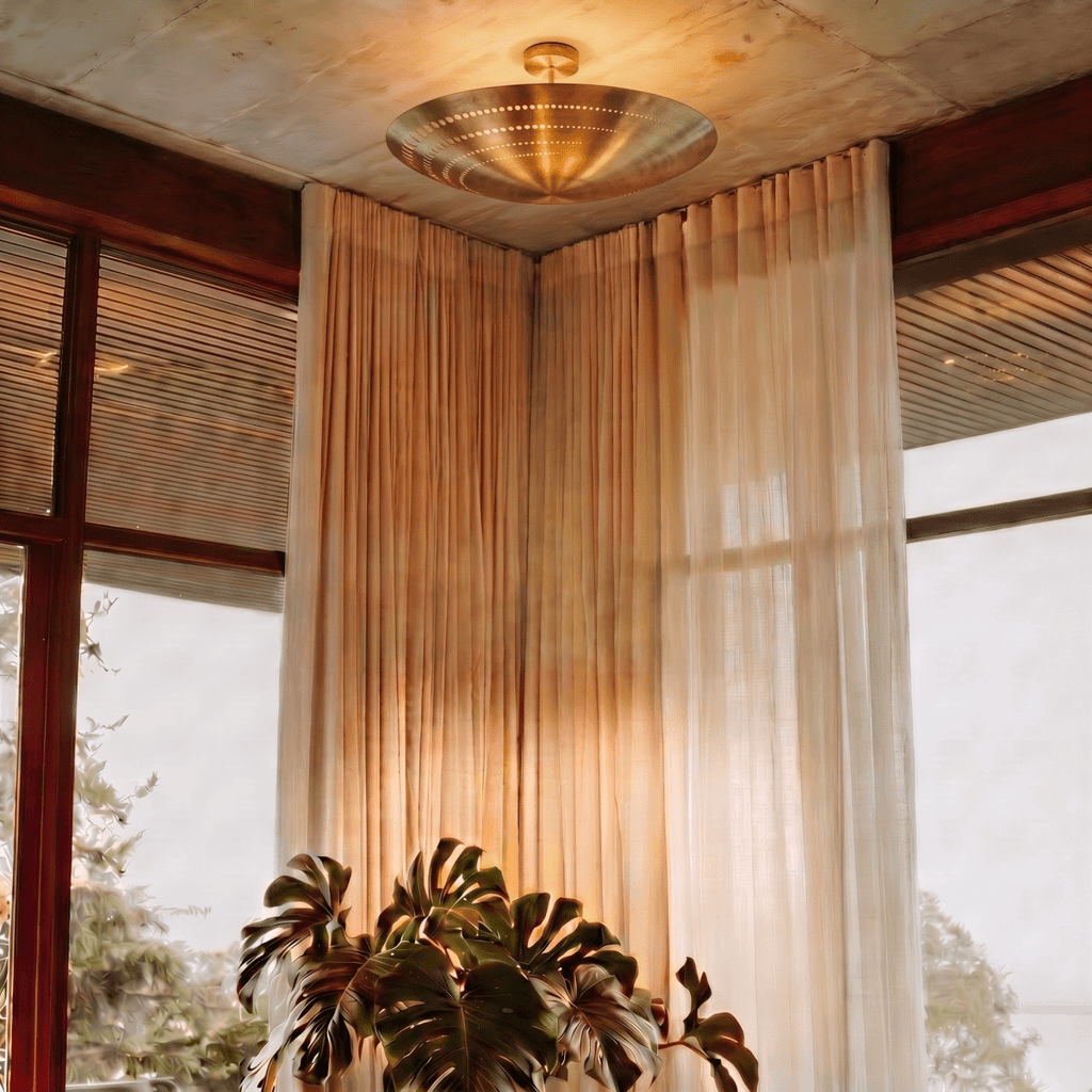 Clérachic ceiling light in brass glowing in a curtained corner beside a large potted monstera plant.