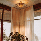 Clérachic ceiling light in brass glowing in a curtained corner beside a large potted monstera plant.