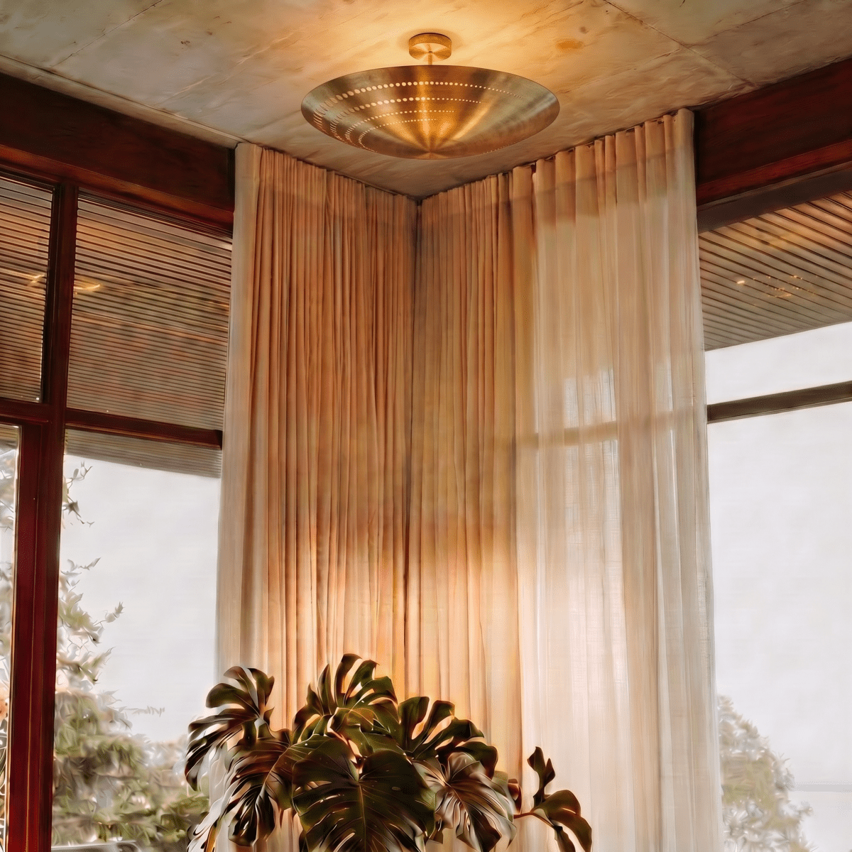 Clérachic ceiling light in brass glowing in a curtained corner beside a large potted monstera plant.