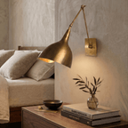 This brass Clérachic wall lamp lights a bedroom nightstand stacked with books beside a small glass vase.