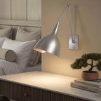 This chrome Clérachic wall lamp lights a marble-top nightstand holding a bonsai and stacked books.