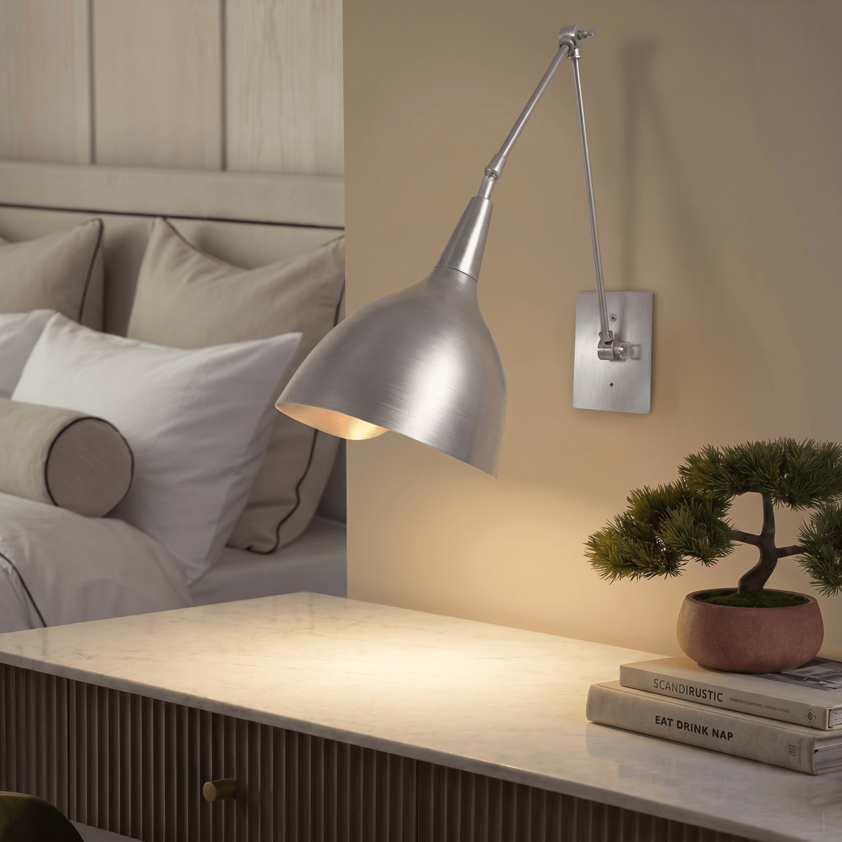 This chrome Clérachic wall lamp lights a marble-top nightstand holding a bonsai and stacked books.