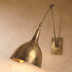 Clérachic wall lamp in vintage brass with a cone-shaped shade on an articulated swing arm against a plaster wall.