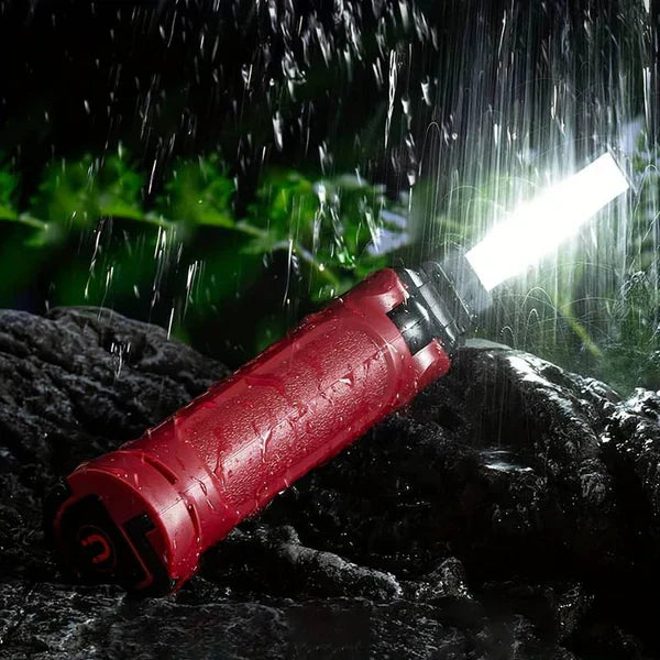 Clérachic work light lying on wet rock in the rain, its COB panel lit against dark foliage.
