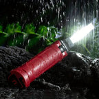 Clérachic work light lying on wet rock in the rain, its COB panel lit against dark foliage.