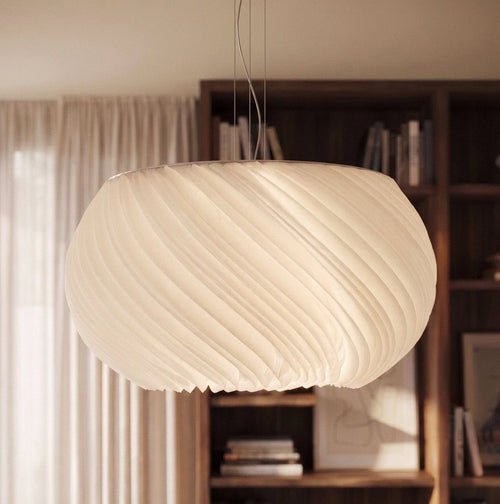 Clérachic pendant shade seen close up, its spiral fluting and scalloped hem in front of a shelf of books and a curtained window.