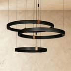 Clérachic ring chandelier in three tiers, black perforated rings stacked with brass mounts against a plaster wall.