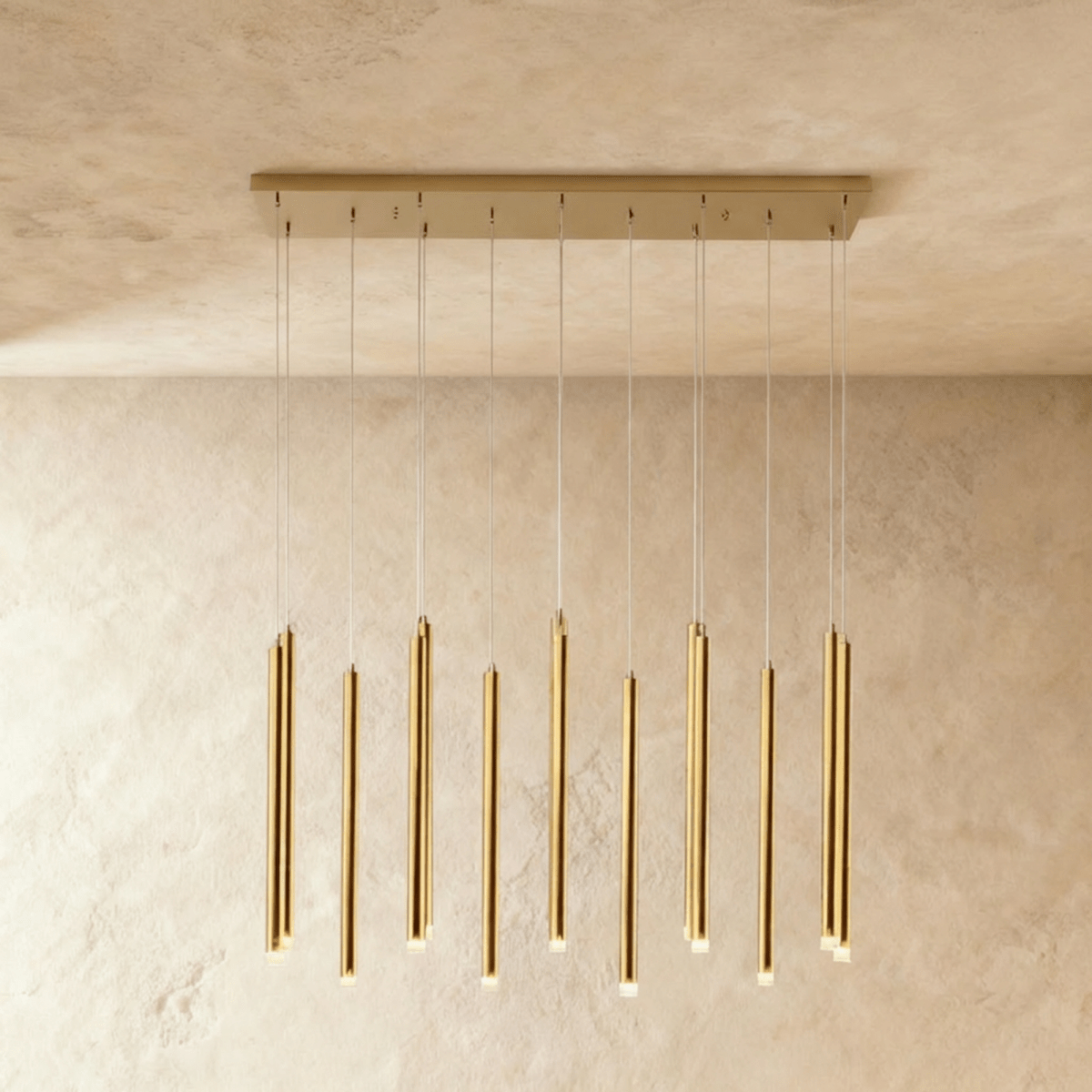 Clérachic gold linear chandelier with slim brass-toned tubes hanging on fine wires from a rectangular canopy, against limewash.