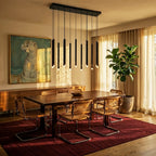 Clérachic black tube chandelier hanging over a walnut dining table with cane chairs, a red rug and a fiddle-leaf fig by sheer curtains.