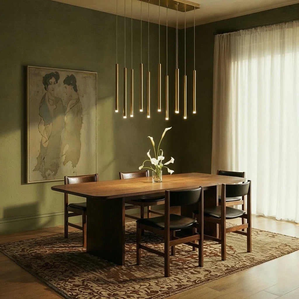 Clérachic gold tube chandelier lit above a long walnut dining table in a green room with calla lilies and black leather chairs.