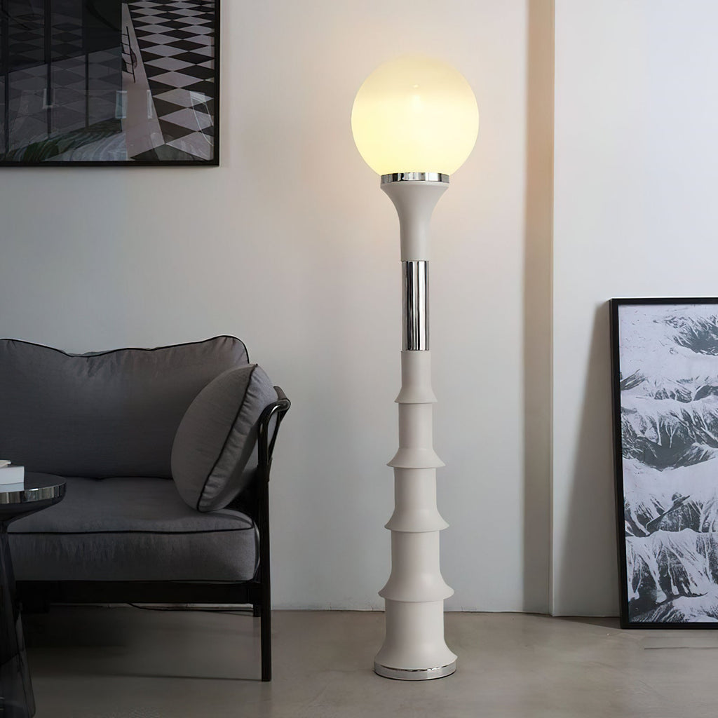 Glowing white globe on a cream, ribbed column with a chrome band, a Clérachic floor lamp beside a grey sofa and a framed print.