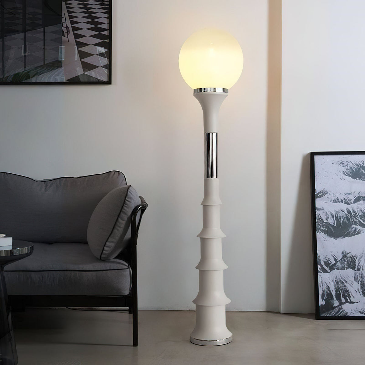 Glowing white globe on a cream, ribbed column with a chrome band, a Clérachic floor lamp beside a grey sofa and a framed print.