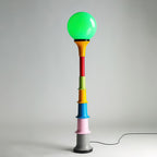 Green-globe Clérachic floor lamp with a yellow funnel, red, green, yellow, blue and pink tiers and a grey foot, on a pale grey backdrop.