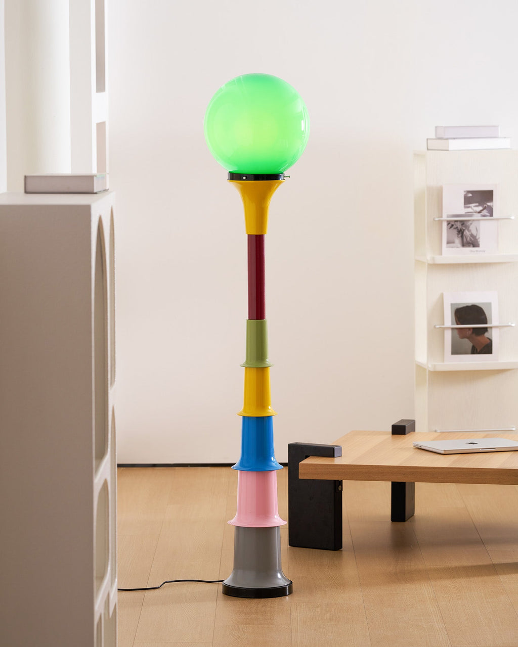 Close crop of a Clérachic floor lamp with a lit green globe and coloured tiers, beside a light wooden table and white shelving.