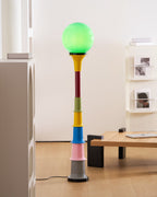Close crop of a Clérachic floor lamp with a lit green globe and coloured tiers, beside a light wooden table and white shelving.