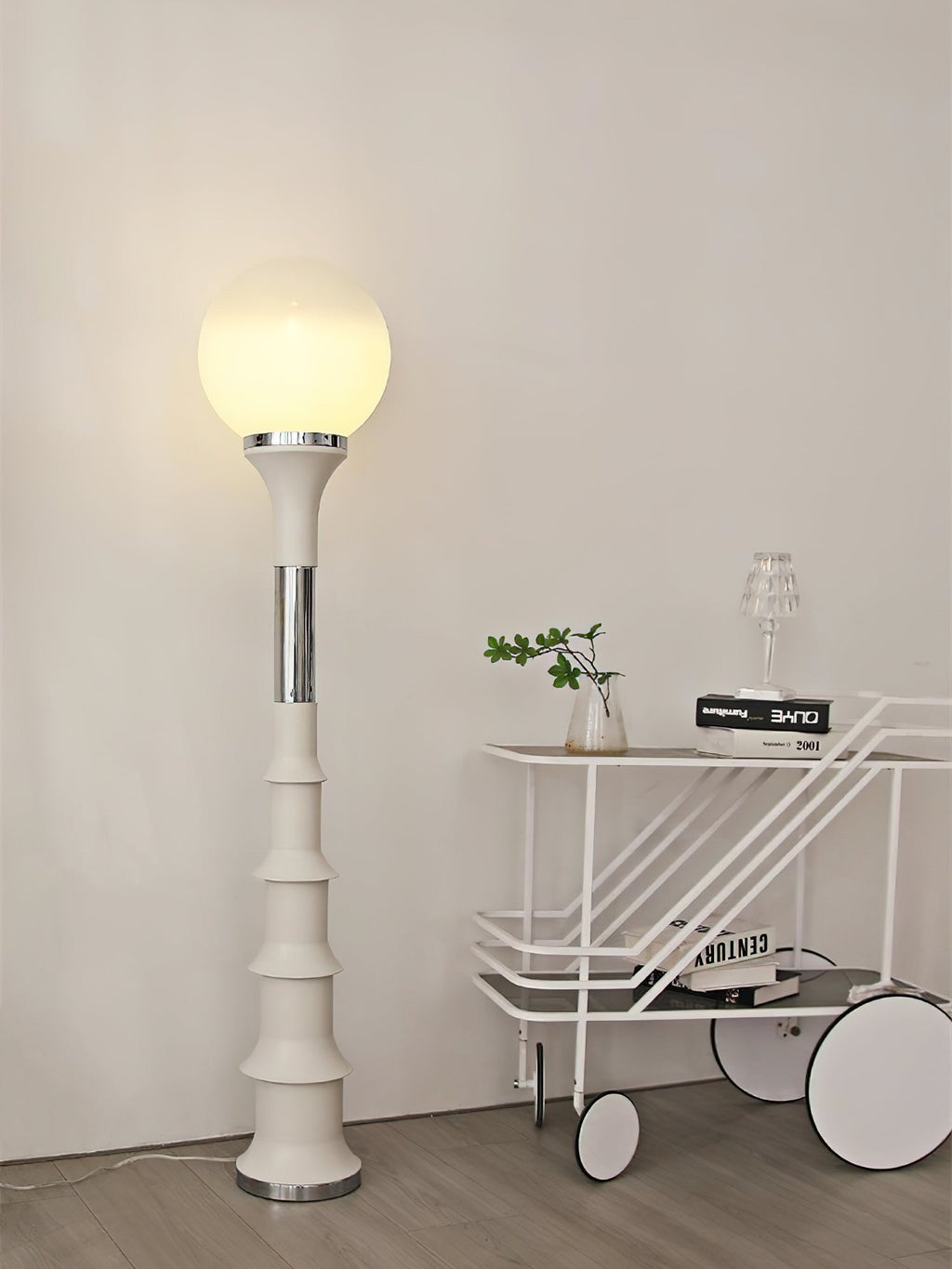 Cream ribbed Clérachic floor lamp with a glowing globe, next to a white trolley with a small plant on it.