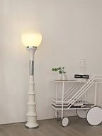 Cream ribbed Clérachic floor lamp with a glowing globe, next to a white trolley with a small plant on it.