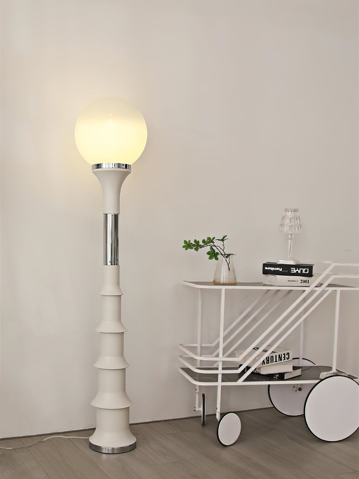 Cream ribbed Clérachic floor lamp with a glowing globe, next to a white trolley with a small plant on it.