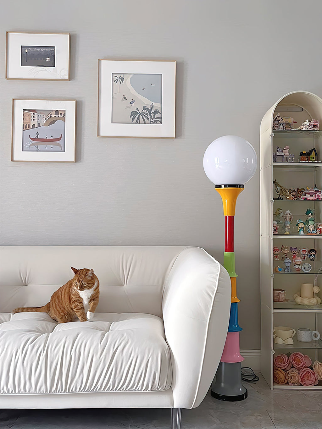 A Clérachic floor lamp with a white globe and multicolour stacked stem, beside a cream sofa with a cat and a display shelf.