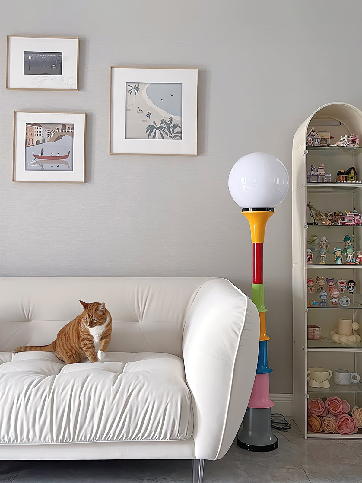 A Clérachic floor lamp with a white globe and multicolour stacked stem, beside a cream sofa with a cat and a display shelf.