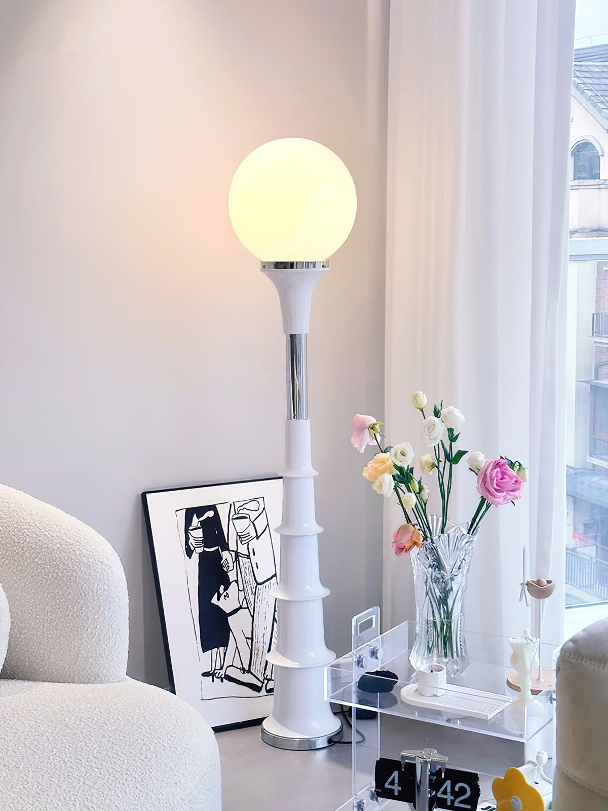 Lit white globe on a cream ribbed stem, a Clérachic floor lamp beside a wall print and a vase of pink and white flowers.
