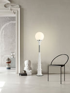 White-globe Clérachic floor lamp on a pale ribbed stem, standing in a large grey room with a black chair and a plaster bust.