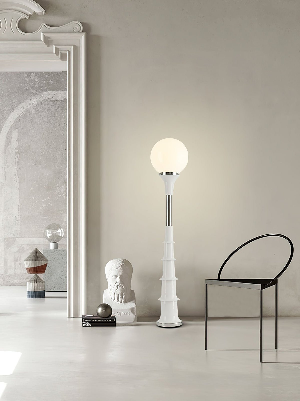 White-globe Clérachic floor lamp on a pale ribbed stem, standing in a large grey room with a black chair and a plaster bust.