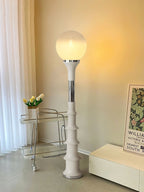 Warm light from the globe of this Clérachic floor lamp, a cream ribbed stem beside a trolley, a print and a window curtain.